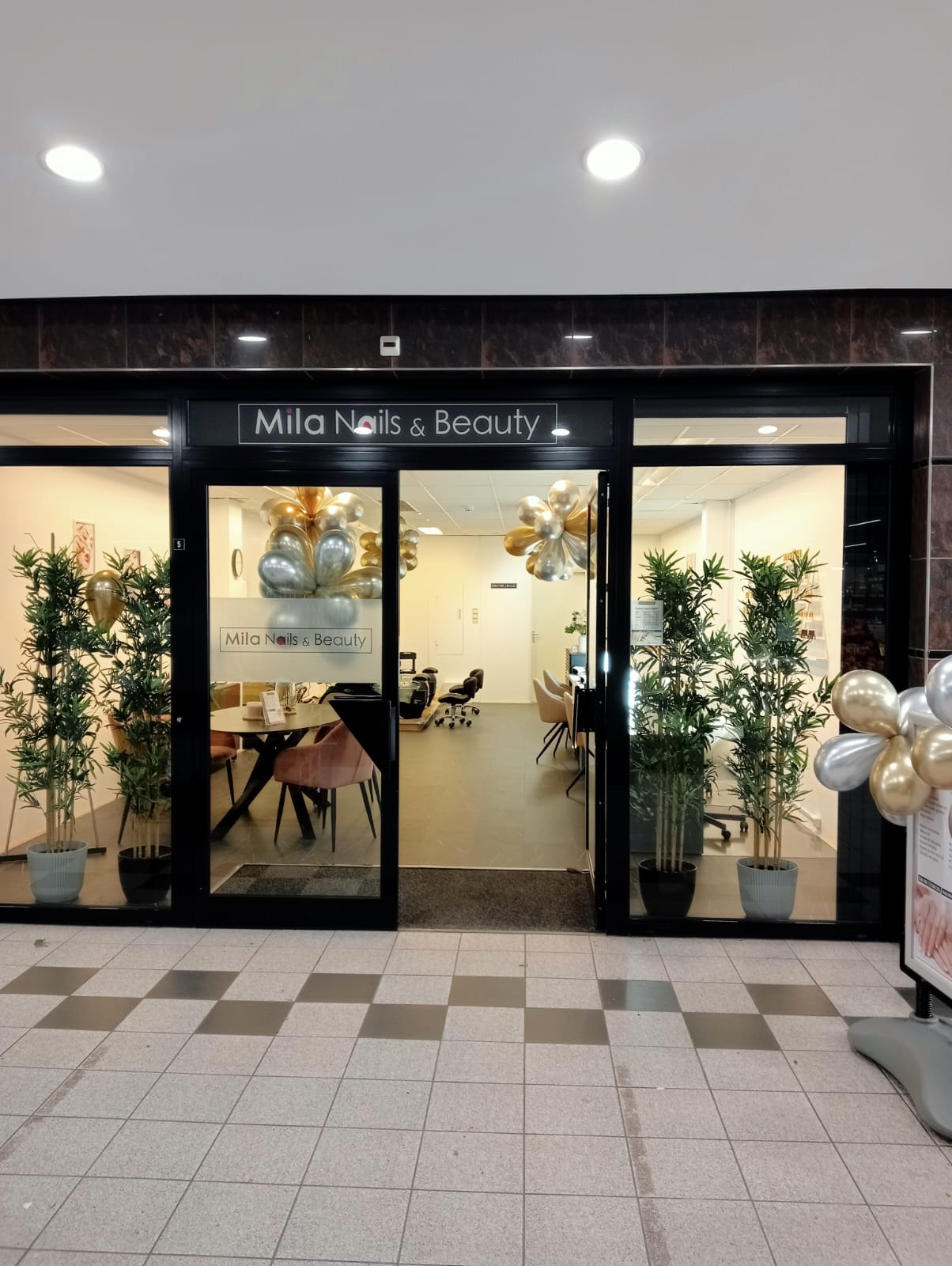 De winkelpui van Mila Nails & Beauty: het naambord boven de glazen deur, planten en gouden en zilveren ballonnen